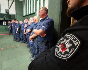 W strzelnicy pod ścianą stoją policjanci, są w tle. Na pierwszym planie widać ramię z naszywką i napis: Policja instruktor strzelań policyjnych.