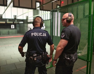 Policjant stoi na lini startu przed tarczą, jest gotowy do strzelania, za nim stoi instruktor.