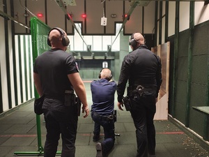 Policjant mierzy do tarczy, klęczy na jedno kolano, za nim stoją dwaj instruktorzy.