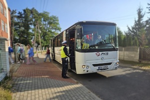 Z autobusu wysiadają dzieci, idą w kierunku szkoły. A przed wejściem do autobusu stoi policjant i rozmawia z kierowcą.
