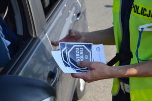 Policjant trzyma w dłoni naklejki i ulotki kampanii &quot;Mistrzu zwolnij&quot;. Policjant stoi obok samochodu.