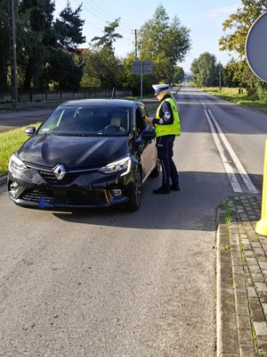 Droga. Policjantka stoi przy samochodzie, od strony kierowcy. Podaje kierującemu urządzenie do badania stanu trzeźwości.