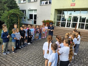 Przed wejściem do szkoły, stoją dzieci, między nimi jest policjantka, mówi do dzieci.