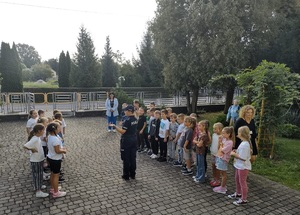 Przed wejściem do szkoły, stoją dzieci, między nimi jest policjantka, mówi do dzieci.