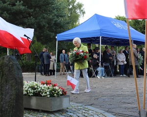 Starsza kobieta idzie pod pomnik z kwiatami by je złożyć.