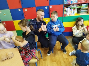 Policjant rozmawia z chłopcem, kuca obok niego, pokazuje mu swoje dłonie.