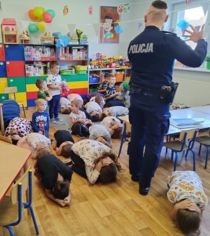 Policjant stoi tyłem do zdjęcia, przed nim na podłodze dzieci leżą w pozycji &quot;na żółwia&quot;, ćwiczą jak zachować się w przypadku ataku agresywnego psa.