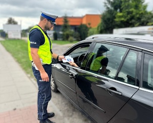 Policjant stoi przy samochodzie od strony kierowcy, wręcza mu ulotkę kampanii &quot;Mistrzu zwolnij&quot;.