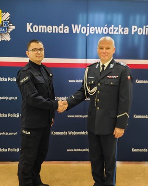 Na tle napisu Komenda Wojewódzka Policji, pozuje do zdjęcia dwóch policjantów. Patrzą w obiektyw, podają sobie ręce.