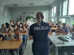 Policjantka stoi tyłem do zdjęcia, przed nią w ławkach siedzą uczniowie.