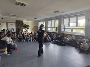 Korytarz w szkole, młodzież siedzi na podłodze wokół policjantki, która stoi i mówi do nich.