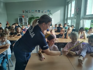 Policjantka rozdaje dzieciom opaski odblaskowe. dzieci siedzą w ławkach szkolnych. Wyciągają ręce po odblask.