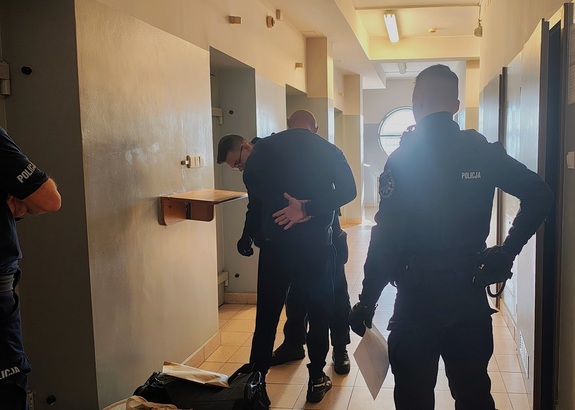 Pomieszczenie PDOZ w komendzie. Policjanci sprawdzają osobę zatrzymaną. Przygotowują do konwoju.
