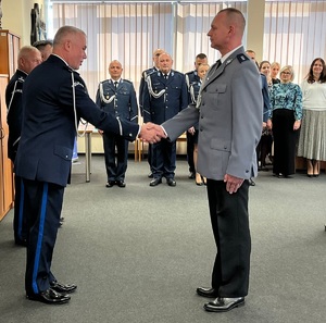 Aula w budynku komendy. Dwaj policjanci podają sobie ręce. Policjant składa meldunek komendantowi.
