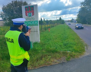 Umundurowany policjant, ma założoną kamizelkę z napisem Policja, patrzy przez urządzenie do mierzenia prędkości jadących samochodów.