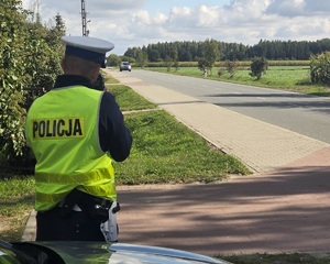 Umundurowany policjant, ma założoną kamizelkę z napisem Policja, patrzy przez urządzenie do mierzenia prędkości jadących samochodów.