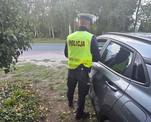 Umundurowany policjant, ma założoną kamizelkę z napisem Policja, stoi przy samochodzie od strony kierowcy, robi kontrolę drogową.