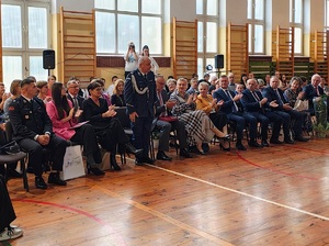 Sala gimnastyczna w szkole, na krzesełkach siedzą goście zaproszeni na uroczystość.