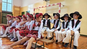W dwóch rzędach ławek siedzą dzieci przebrane w stroje góralskie. W pierwszym rzędzie siedzą dziewczynki, a w drugim chłopcy.