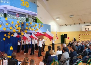 Sala gimnastyczna w szkole. W tle scenografia - duże drzewo z kwiatami. Na ich tle tańczą dziewczynki, każda z nich trzyma w dłoni polską flagę narodową.