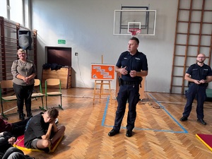 Sala gimnastyczna w szkole. Na środku stoją dwaj policjanci i kobieta. Z tyłu za policjantem tablica stojąca na sztaludze z napisem: Tydzień Przeciwko Przemocy Rówieśniczej.