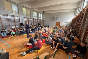 W sali gimnastycznej, na materacach na podłodze siedzą uczniowie. Przed nimi stoi policjantka, mówi do nich.