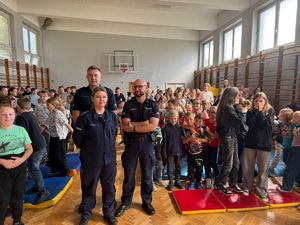 Dwóch policjantów i policjantka pozują do zdjęcia na tle młodzieży, na sali gimnastycznej.