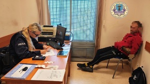 Pokój w komendzie, po prawej stronie na krzesełku siedzi mężczyzna, po lewej za biurkiem siedzi policjantka, uzupełnia dokumentację.