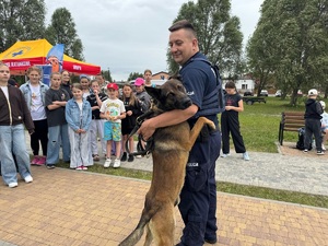 Pies przytula się do policjanta swojego przewodnika. W tle stoją dzieci i się przyglądają.
