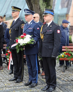 Umundurowani funkcjonariusze Policji, Straży Pożarnej i Straży Miejskiej. Policjant trzyma w dłoniach kwiaty.