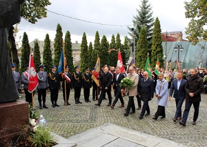 Samorządowcy składają kwiaty pod pomnikiem św. Maksymiliana. W tle poczty sztandarowe służb mundurowych i szkół.