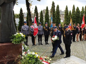 Funkcjonariusze składają kwiaty pod pomnikiem. W tle poczty sztandarowe służb mundurowych i szkół.