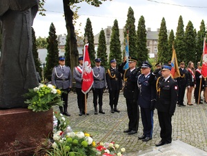 Funkcjonariusze salutuja  pod pomnikiem. W tle poczty sztandarowe służb mundurowych i szkół.