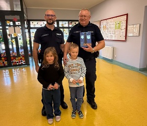 Dwóch policjantów pozuje do zdjęcia z chłopcem i dziewczynką. Policjant trzyma w ręku laurkę od dzieci.
