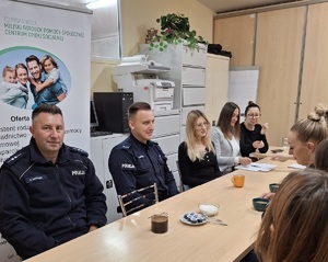 Policjanci i pracownicy socjalni siedzą wspólnie przy stole, mają w kubkach picie. Rozmawiają.