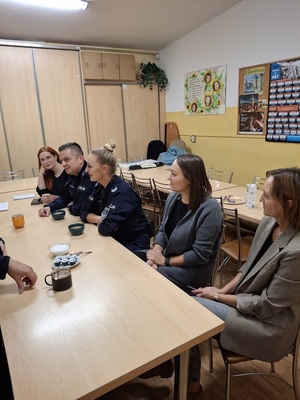Policjanci i pracownicy socjalni siedzą wspólnie przy stole, mają w kubkach picie. Rozmawiają.