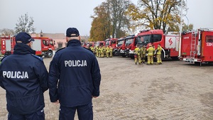 Dwaj policjanci stoją na placu przed zebranymi strażakami i wozami strażackimi.