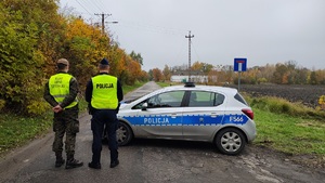 Radiowóz policyjny, przy nim stoi policjant i funkcjonariusz Wojsk Obrony Terytorialnej. Zastawili radiowozem drogę, by nikt nie wjechał na teren ćwiczeń.