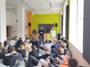 Sala szkolna. Na podłodze są rozłożone poduchy-pufy, na których siedzi młodzież. Na wprost nich stoi policjantka i mówi do zebranych. Obok policjantki stoi nauczycielka.