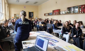 Sala w szkole, uczniowie siedzą na krzesełkach przy dużych stołach. Tylem do zdjęcia siedzi policjantka, mówi do młodzieży, która patrzy na nią i słucha co mówi.