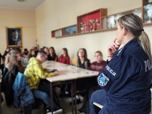 Sala w szkole, uczniowie siedzą na krzesełkach przy dużych stołach. Tylem do zdjęcia siedzi policjantka, mówi do młodzieży, która patrzy na nią i słucha co mówi.