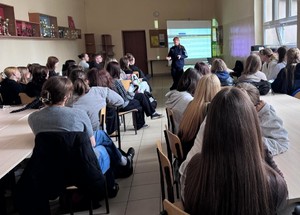 Sala w szkole, uczniowie siedzą na krzesełkach przy dużych stołach. Policjantka, mówi do młodzieży, która patrzy na nią i słucha co mówi.