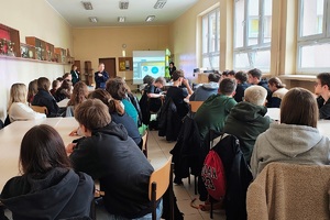 Sala w szkole, uczniowie siedzą na krzesełkach przy dużych stołach. Policjantka, mówi do młodzieży, która patrzy na nią i słucha co mówi.