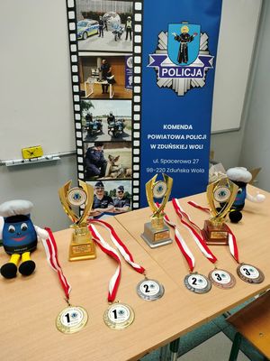 Roll-up Komendy Powiatowej Policji w Zduńskiej Woli na tle którego stoją puchary oraz medale dla laureatów konkursu.