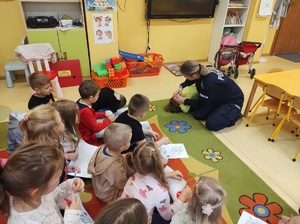 W sali na dywanie siedzą dzieci, na wprost nich policjantka i jedno dziecko pokazują bezpieczną pozycję &quot;na żółwia&quot;.