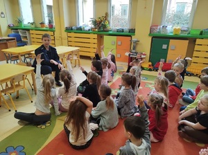 Dzieci siedzą na dywanie, podnoszą do góry rączki, zgłaszają się do odpowiedzi. Policjantka siedzi na wprost nich na krzesełku.