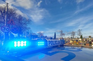 Radiowóz z włączonymi światłami na dachu. W tle kościół i cmentarz.