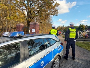 Policjanci stoją przed wejściem na cmentarza. Obok nich stoi radiowóz.
