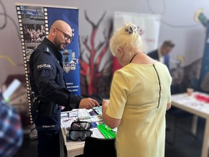 Policjant stoi przy stoisku informacyjnym, obok niego tyłem do zdjęcia stoi kobieta, ogląda co leży na stole.