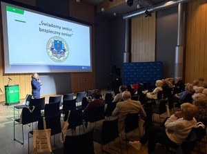 Sala, na krzesełkach, tyłem do zdjęcia siedzi publiczność. Na wprost nich na ścianie jest rzutnik, wyświetlana jest prezentacja. Pod ekranem stoi policjantka, mówi do mikrofonu.